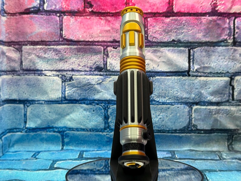 May include: A silver and gold lightsaber prop with black accents, standing upright on a black stand. The lightsaber features a cylindrical design with detailed ridges and a gold emitter. The background is a colorful brick wall.