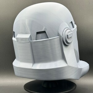 May include: A white plastic helmet with a textured surface and a raised detail on the side. The helmet is on a black stand.