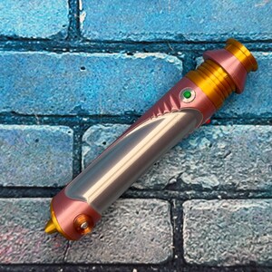 Op de afbeelding: Een roze, gouden en zilveren lichtzwaard prop met een groene knop. Het handvat is roze en goud, met een zilveren bladgedeelte. Het lichtzwaard staat tegen een blauwe bakstenen muur.