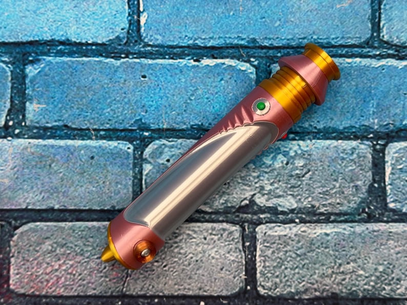 Op de afbeelding: Een roze, gouden en zilveren lichtzwaard prop met een groene knop. Het handvat is roze en goud, met een zilveren bladgedeelte. Het lichtzwaard staat tegen een blauwe bakstenen muur.