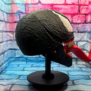 May include: A black Venom mask with a textured surface, white eyes, and a long red tongue. The mask is displayed on a black stand against a brick wall backdrop.