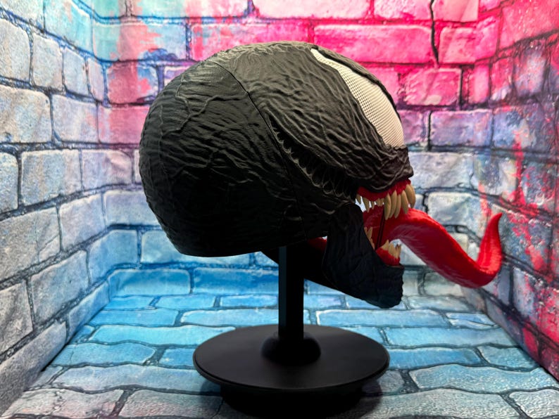 May include: A black Venom mask with a textured surface, white eyes, and a long red tongue. The mask is displayed on a black stand against a brick wall backdrop.