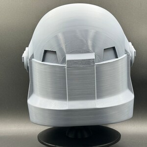 May include: A white 3D printed helmet with a textured surface. The helmet has a rounded top and a flat bottom. The helmet is sitting on a black base.