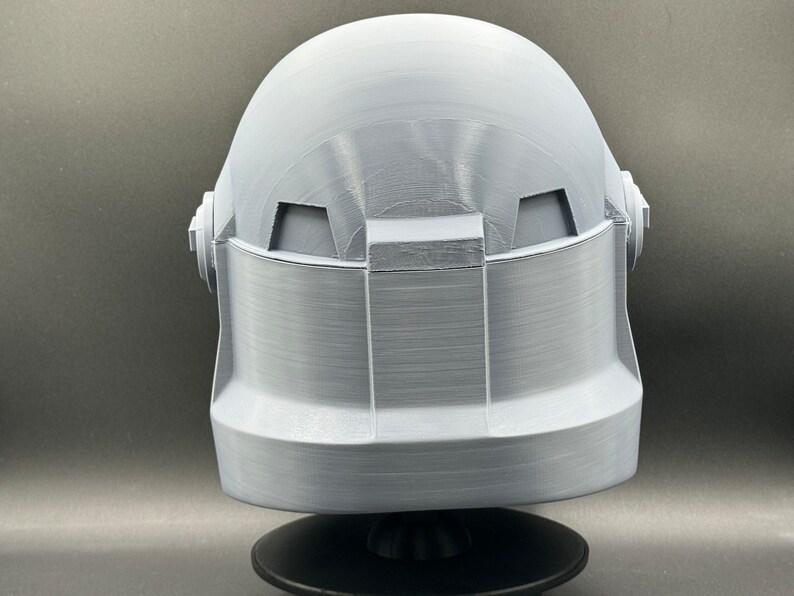 May include: A white 3D printed helmet with a textured surface. The helmet has a rounded top and a flat bottom. The helmet is sitting on a black base.