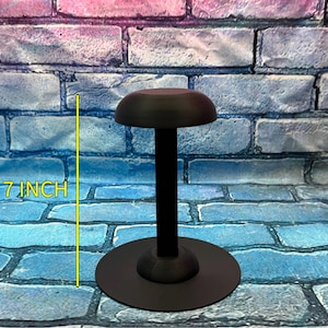 May include: A black hat stand, approximately 7 inches tall, with a round base and a domed top. The stand is made of a matte black material and is designed to display hats. The background is a brick wall.