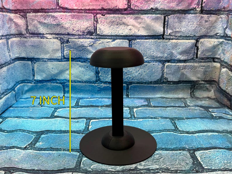 May include: A black hat stand, approximately 7 inches tall, with a round base and a domed top. The stand is made of a matte black material and is designed to display hats. The background is a brick wall.