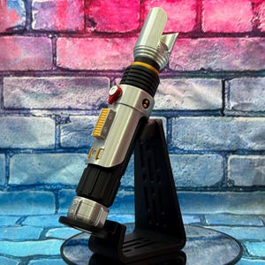 May include: A silver and black lightsaber prop with yellow and red accents, resting on a black stand. The prop has a detailed design with a cylindrical body and a blade emitter. The background is a brick wall with blue, pink, and purple hues.