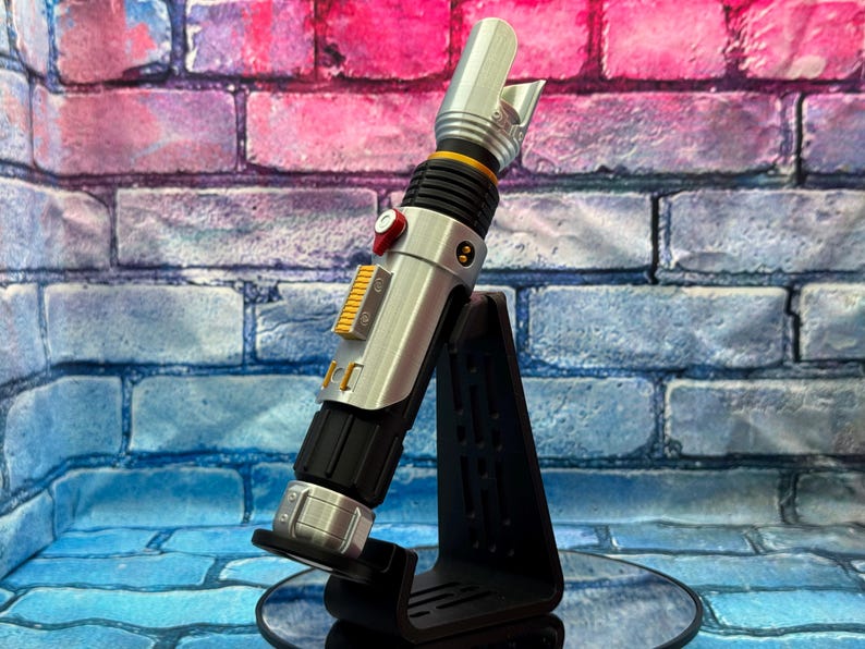 May include: A silver and black lightsaber prop with yellow and red accents, resting on a black stand. The prop has a detailed design with a cylindrical body and a blade emitter. The background is a brick wall with blue, pink, and purple hues.