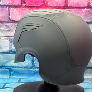 May include: A gray helmet with a futuristic design, featuring angular panels and textured surfaces. The helmet has a visor area and is mounted on a black stand. The overall aesthetic suggests a sci-fi or cosplay theme.