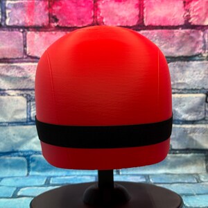 May include: A bright red, smooth, rounded object with a horizontal black elastic band encircling the middle section. The object is mounted on a black circular stand, presenting a clean, uniform surface texture without any additional markings or details.