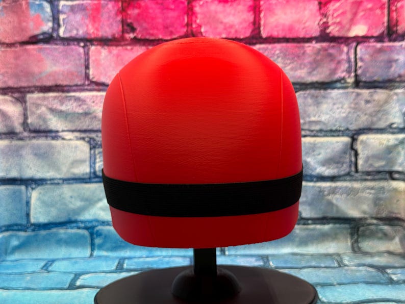 May include: A bright red, smooth, rounded object with a horizontal black elastic band encircling the middle section. The object is mounted on a black circular stand, presenting a clean, uniform surface texture without any additional markings or details.