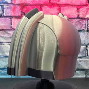 May include: A futuristic-looking helmet with a metallic silver and reddish-brown finish. The helmet has a curved design with black accents and is mounted on a black stand. The background features a colorful brick wall.