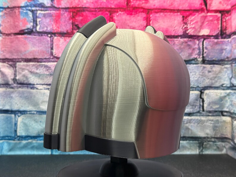 May include: A futuristic-looking helmet with a metallic silver and reddish-brown finish. The helmet has a curved design with black accents and is mounted on a black stand. The background features a colorful brick wall.