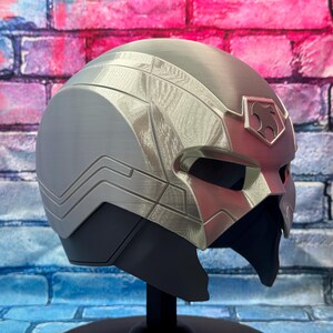 May include: A silver and gray helmet with a sleek, futuristic design. The helmet features angular lines, a visor, and a decorative emblem on the forehead. The color scheme includes shades of silver, gray, and black, with a hint of red. The helmet is displayed on a black stand.