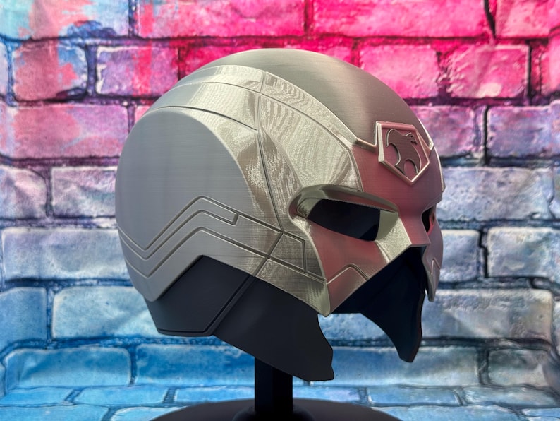 May include: A silver and gray helmet with a sleek, futuristic design. The helmet features angular lines, a visor, and a decorative emblem on the forehead. The color scheme includes shades of silver, gray, and black, with a hint of red. The helmet is displayed on a black stand.