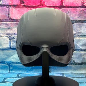 May include: A gray, futuristic-looking helmet with large eye openings, displayed on a black stand. The helmet has a textured surface and a streamlined design, suggesting a sci-fi or cosplay theme. The background is a colorful brick wall.