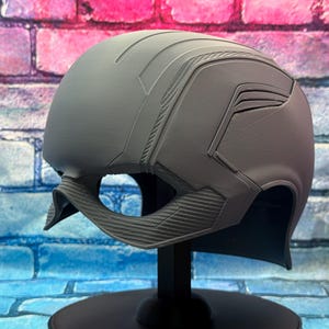 May include: A gray helmet with a detailed design, featuring ridges and angular panels. The helmet is displayed on a black stand against a colorful brick wall background. The helmet's design includes a chin strap.