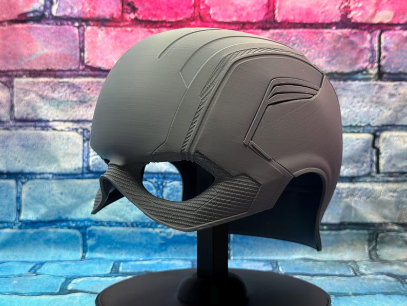 May include: A gray helmet with a detailed design, featuring ridges and angular panels. The helmet is displayed on a black stand against a colorful brick wall background. The helmet's design includes a chin strap.