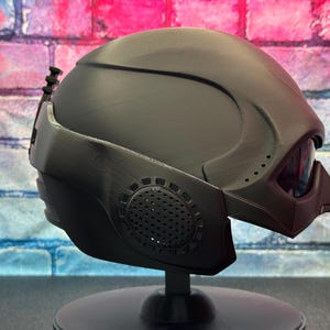 May include: A matte black helmet with a futuristic design, featuring a visor and integrated details. The helmet is mounted on a black stand, showcasing its angular shape and textured surfaces. The design includes vents and a distinct, pointed visor.