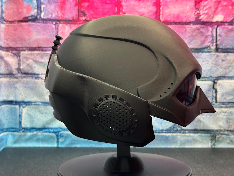 May include: A matte black helmet with a futuristic design, featuring a visor and integrated details. The helmet is mounted on a black stand, showcasing its angular shape and textured surfaces. The design includes vents and a distinct, pointed visor.