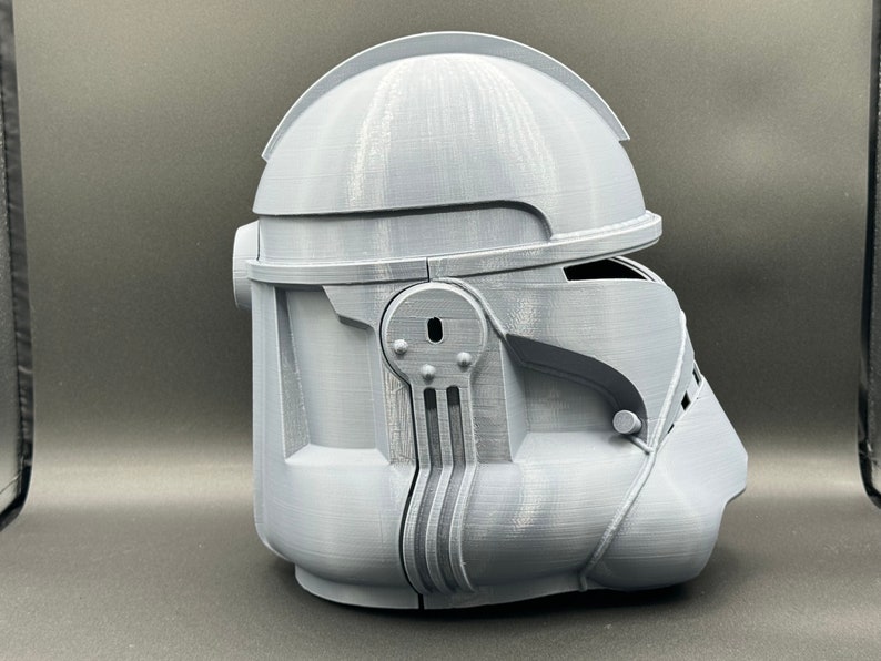 Puede incluir: Un casco gris impreso en 3D con un dise&ntilde;o detallado. El casco tiene una visera y una superficie texturizada.