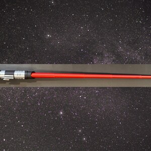 May include: A red lightsaber with a silver and black hilt. The lightsaber is displayed horizontally against a starry background. The lightsaber is a prop from the Star Wars franchise.