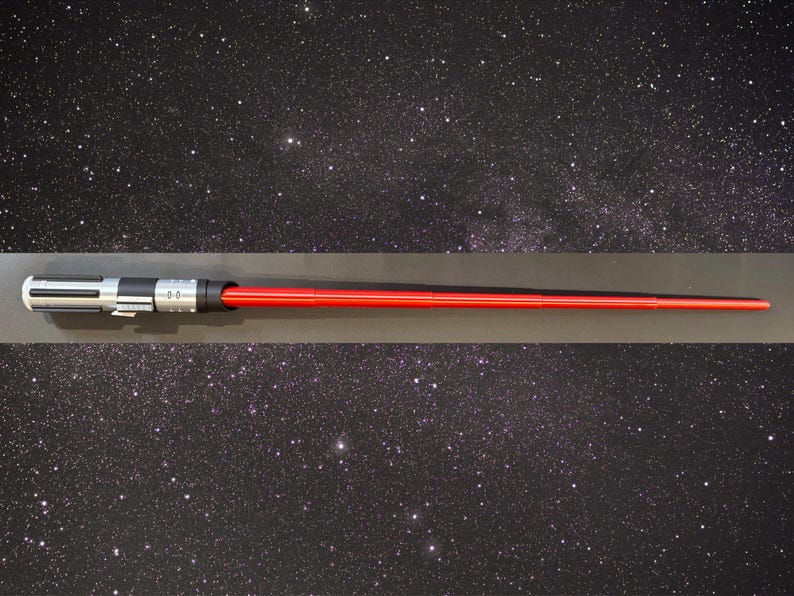 May include: A red lightsaber with a silver and black hilt. The lightsaber is displayed horizontally against a starry background. The lightsaber is a prop from the Star Wars franchise.