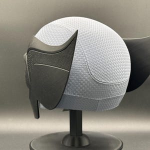 May include: A black and gray 3D printed helmet with a textured surface. The helmet has a pointed ear on the right side and a large, pointed ear on the left side.