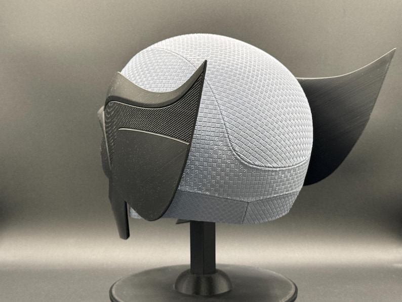 May include: A black and gray 3D printed helmet with a textured surface. The helmet has a pointed ear on the right side and a large, pointed ear on the left side.