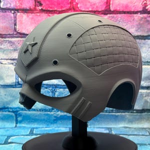 May include: A gray helmet with a star emblem and a woven pattern on the back. The helmet has eye openings and is displayed on a black stand. The background is a brick wall.