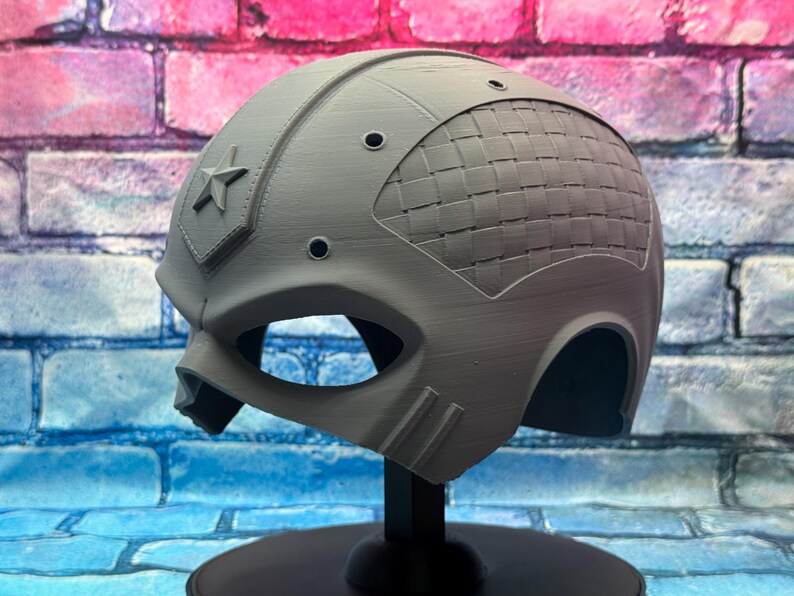 May include: A gray helmet with a star emblem and a woven pattern on the back. The helmet has eye openings and is displayed on a black stand. The background is a brick wall.