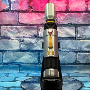 May include: A silver and black lightsaber prop with yellow and red accents. The prop is displayed on a black stand against a brick wall background. The lightsaber has a cylindrical design with detailed features.