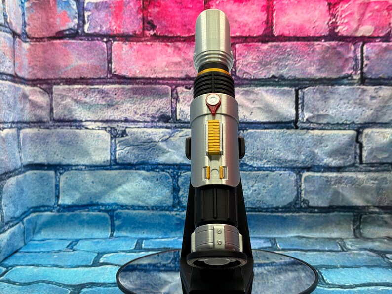 May include: A silver and black lightsaber prop with yellow and red accents. The prop is displayed on a black stand against a brick wall background. The lightsaber has a cylindrical design with detailed features.