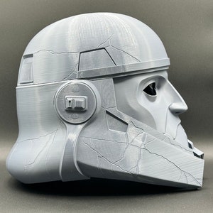 May include: A gray 3D printed helmet with a textured surface and a visor. The helmet has a detailed design with a raised ridge on the top and a small, round detail on the side.