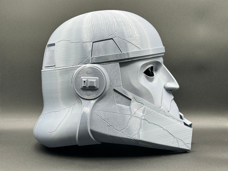 May include: A gray 3D printed helmet with a textured surface and a visor. The helmet has a detailed design with a raised ridge on the top and a small, round detail on the side.