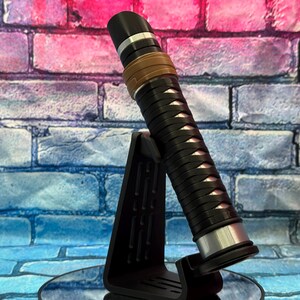 May include: A black and silver lightsaber with a brown grip and a black stand. The lightsaber has a detailed design with silver accents and a black handle. The background is a colorful brick wall.