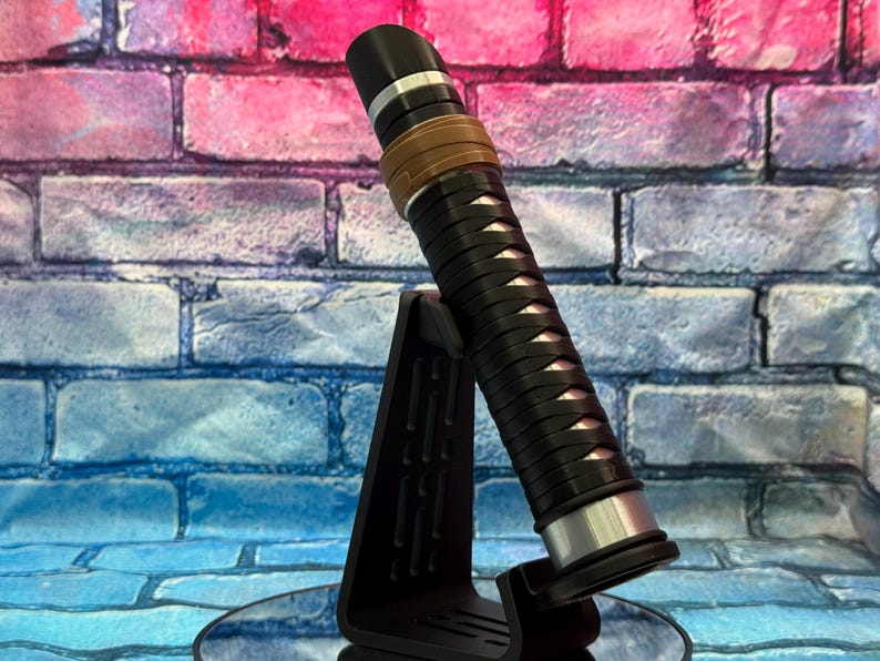 May include: A black and silver lightsaber with a brown grip and a black stand. The lightsaber has a detailed design with silver accents and a black handle. The background is a colorful brick wall.