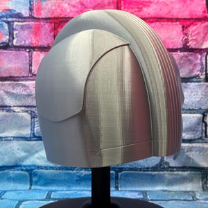 May include: A light gray helmet with a curved visor and ridged detailing on the top. The helmet is displayed on a black stand against a brick wall background.