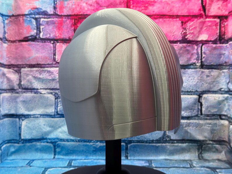 May include: A light gray helmet with a curved visor and ridged detailing on the top. The helmet is displayed on a black stand against a brick wall background.