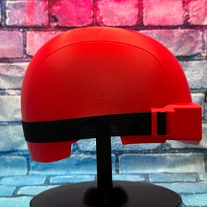 May include: A bright red, smooth-surfaced helmet featuring a rounded dome shape and a prominent rectangular protrusion on the side. A black elastic strap wraps horizontally around the base of the helmet, secured with a black plastic buckle.