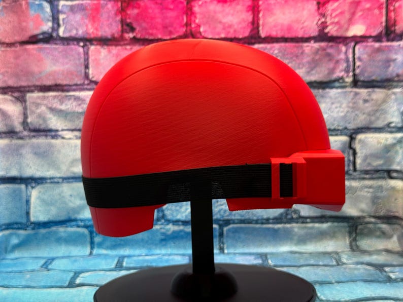 May include: A bright red, smooth-surfaced helmet featuring a rounded dome shape and a prominent rectangular protrusion on the side. A black elastic strap wraps horizontally around the base of the helmet, secured with a black plastic buckle.