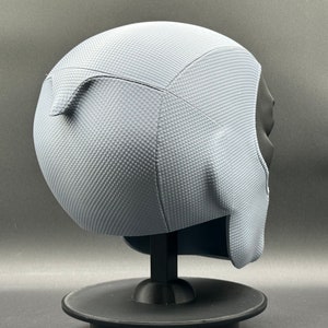 Może przedstawiać: Szary kask z fakturowaną powierzchnią. Kask stoi na czarnym stojaku.