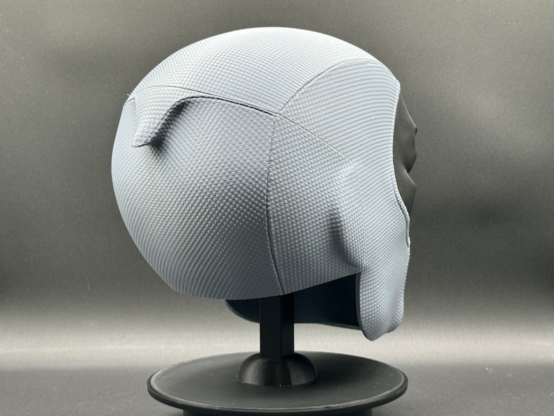 Może przedstawiać: Szary kask z fakturowaną powierzchnią. Kask stoi na czarnym stojaku.