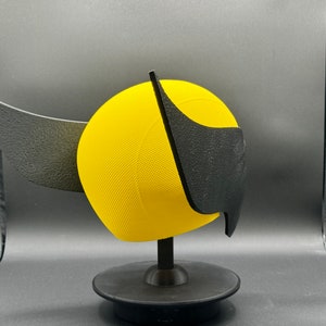 Puede incluir: Un casco impreso en 3D amarillo y negro con un dise&ntilde;o de ala. El casco est&aacute; en un soporte negro.