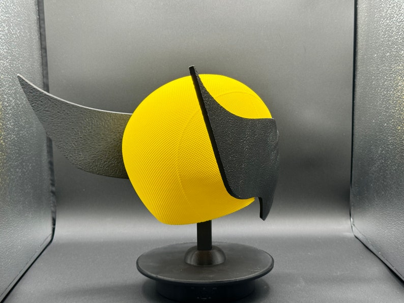Puede incluir: Un casco impreso en 3D amarillo y negro con un dise&ntilde;o de ala. El casco est&aacute; en un soporte negro.