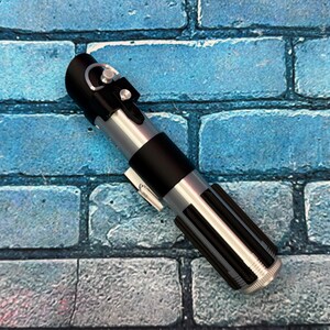 May include: A replica lightsaber with a black handle, silver accents, and a ribbed lower section. The prop is set against a blue brick wall background. The lightsaber is a detailed representation of a popular science fiction weapon.