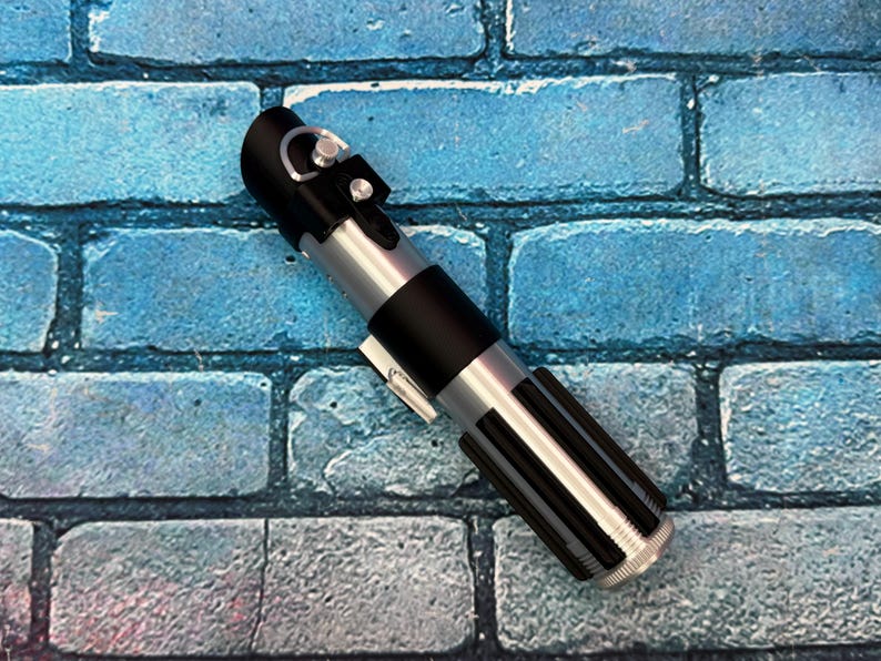 May include: A replica lightsaber with a black handle, silver accents, and a ribbed lower section. The prop is set against a blue brick wall background. The lightsaber is a detailed representation of a popular science fiction weapon.