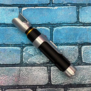 May include: A silver and black lightsaber prop with a gold accent ring. The hilt is black with a ribbed texture, and the emitter is silver. The lightsaber is set against a blue brick wall.