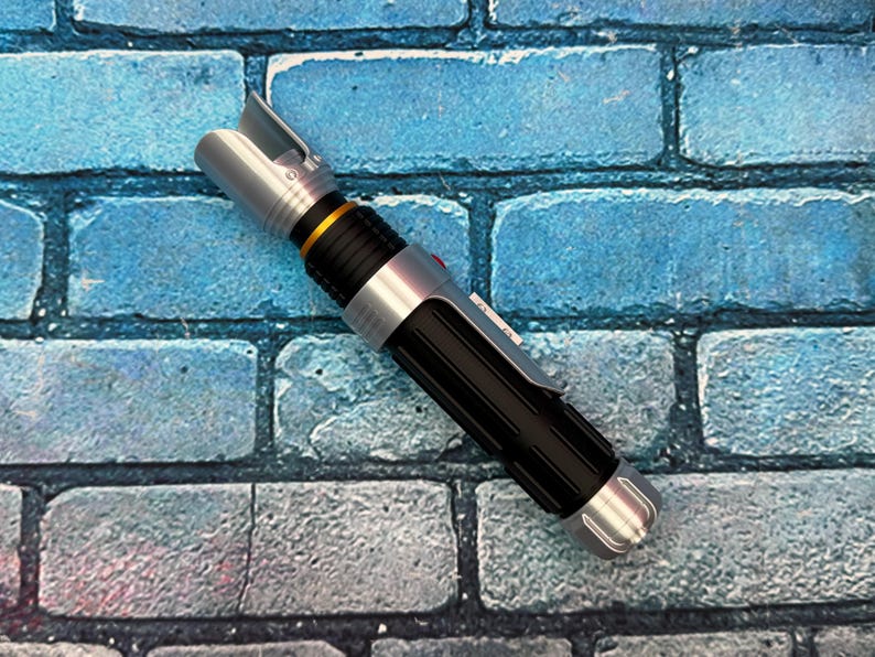 May include: A silver and black lightsaber prop with a gold accent ring. The hilt is black with a ribbed texture, and the emitter is silver. The lightsaber is set against a blue brick wall.