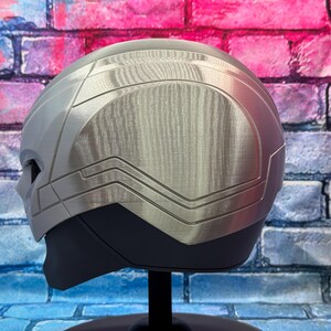May include: A silver and gray helmet with a textured metallic finish. The helmet has a streamlined design with angular details and a dark gray lower section. It is displayed on a black stand against a brick wall.